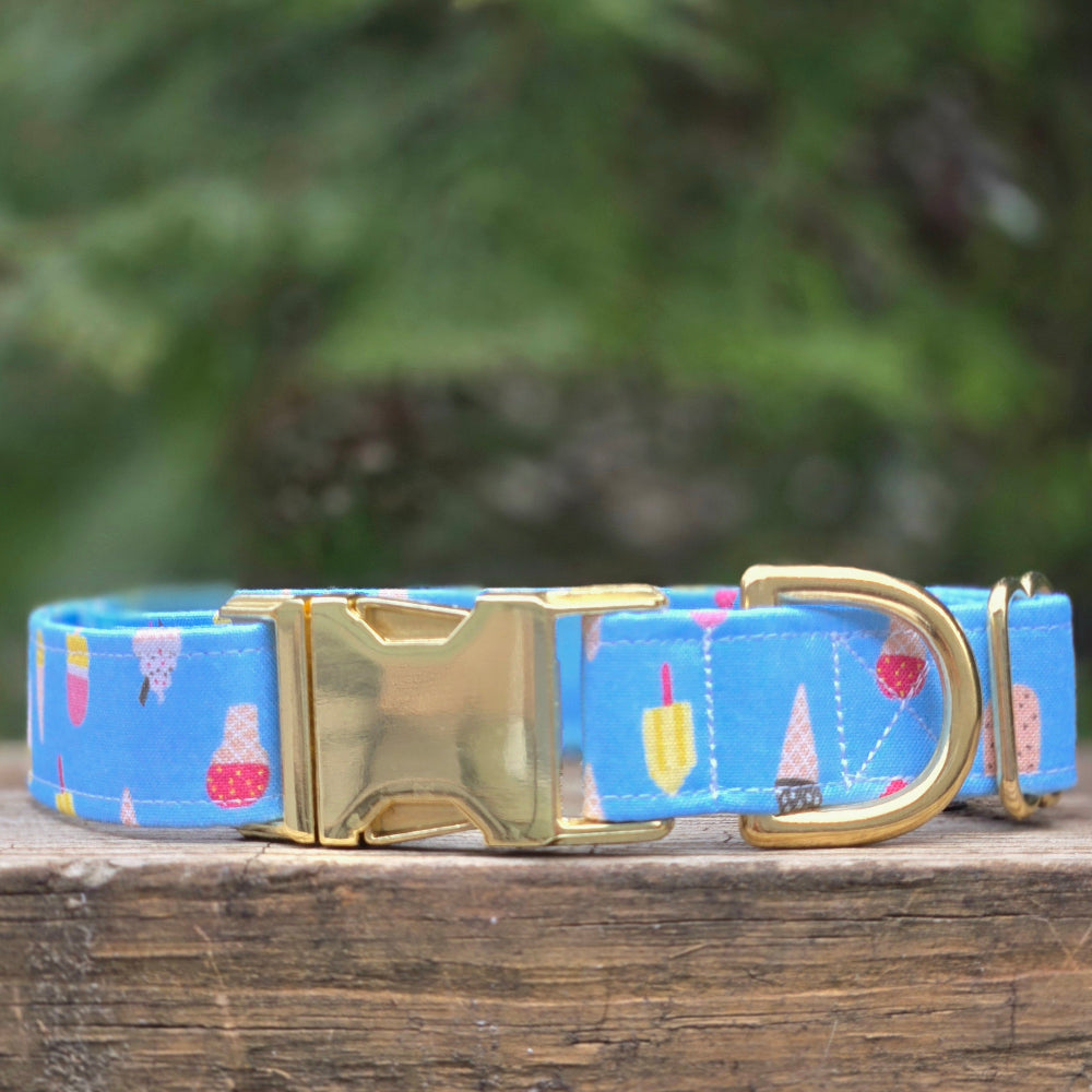 Dog collar with pastel assorted popsicles on a light blue background and gold buckle. on a wooden surface with blurred greenery in the background