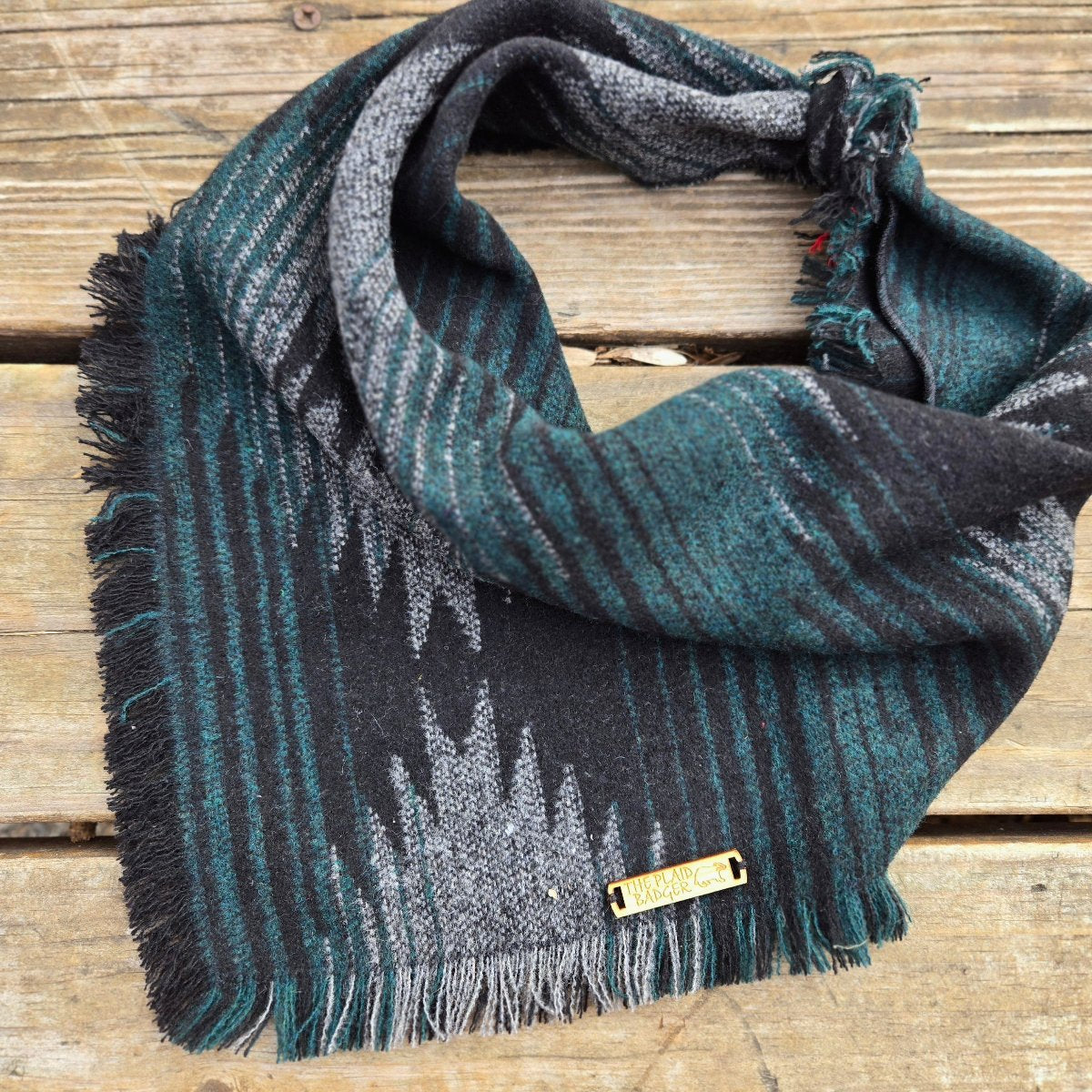 A heavyweight knit dog bandana in a southwest, Aztec style pattern. Shades of gray and emerald green on a black background. Edges have been lightly frayed.