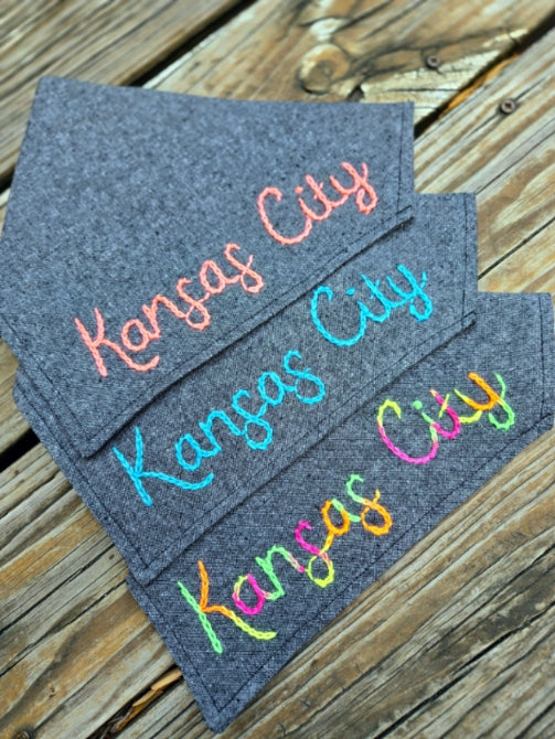 Kansas City dog bandana made from a heathered black cotton linen blend. Bandanas are over the collar in style. Kansas City has been lovingly hand lettered in a cursive type script and hand embroidered in your choice of thread. Shown with neon watermelon, arctic blue, and neon rainbow. Bright and high contrast.