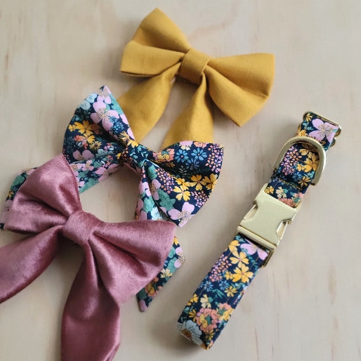 wildflower dog collar, a bright variety of flowers inspired by the 1970s, on a dark background. shown with matching fancy girl dog bow in the same pattern as well as coordinating bows, our mustard linen fancy girl bow and our rose gold velvet fancy girl dog bow  (bows sold separately)