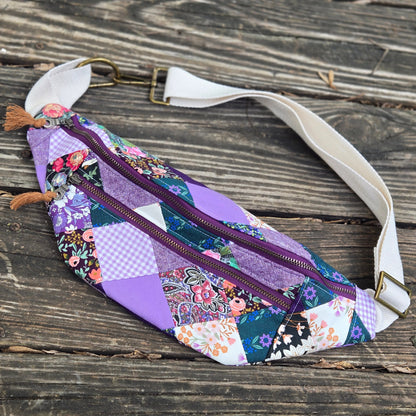 A patchwork fabric bag with purple, floral, and gingham patterns, featuring a white adjustable strap, metal buckle, and brown tassels, displayed on a rustic wooden surface.
