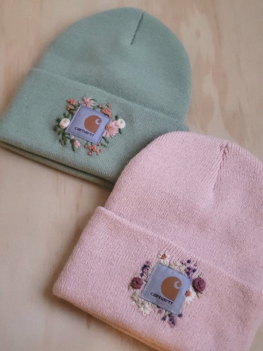 warm, cozy, carhartt brand acrylic knit winter beanie hat, one size fits all available in multi colors, but shown in mint green, and light dusty rose pink. beanies have been hand embroidered around carhartt logo patch with a variety of embroidery flosses with hand embroidered, roses, daisies, leaves, and other flowers in pretty complementary colors to their hat. 